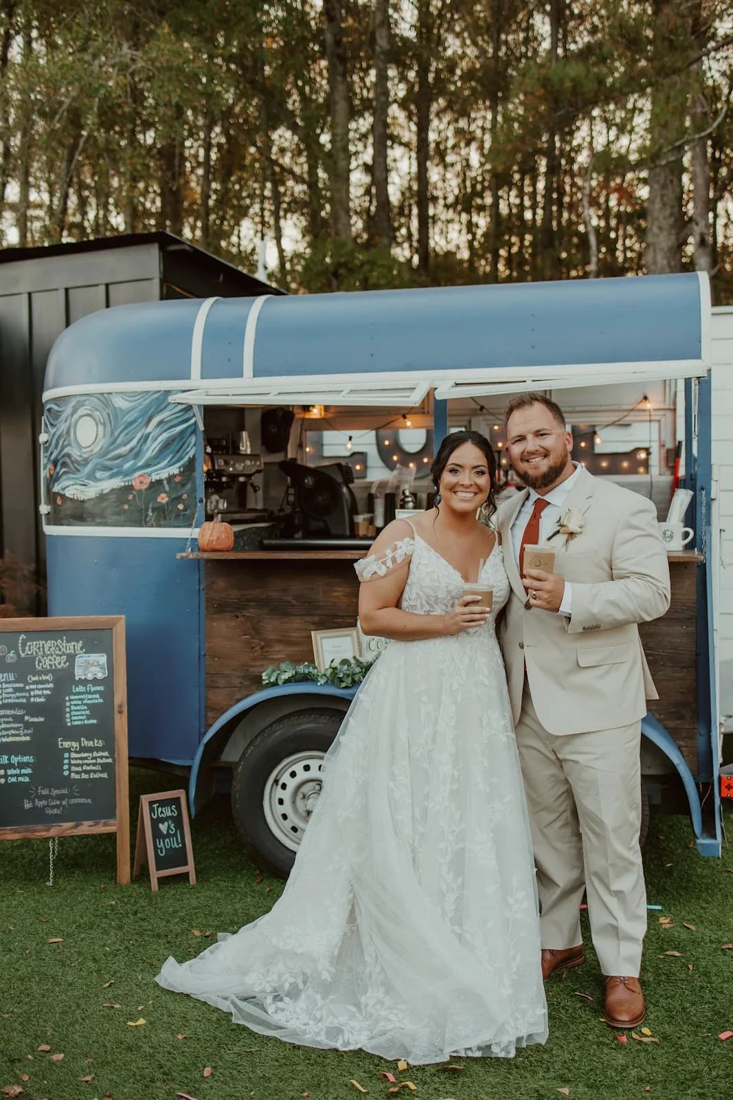 A bride and groom holding iced coffees in front of Cornerstone Coffee's mobile coffee trailer and chalkboard menu