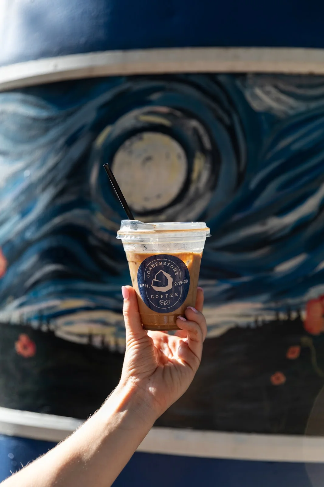 A branded Cornerstone Coffee cup held up in front of the trailer's hand-painted starry mural